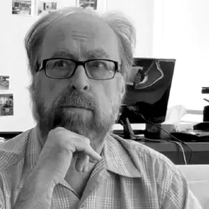 A bearded man wearing glasses is sitting at a desk, thoughtfully resting his chin on his hand. Behind him is a computer monitor and a whiteboard with some photos or papers attached. The image is in black and white.