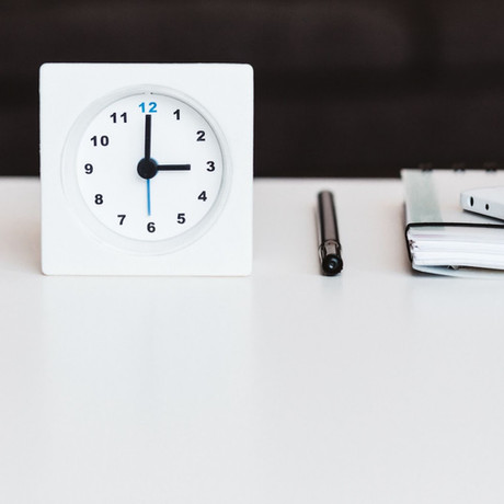 A modern white desk clock showing the time at 10:10, next to a pen and a stack of notebooks, symbolizing time management.