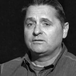 A man with short hair in a denim shirt looks slightly off-camera. The background is dark, and the image is in black and white.