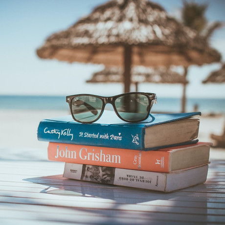 A stack of books and sunglasses on a beach table, with palm trees and the ocean in the background, representing relaxation and vacation reading.