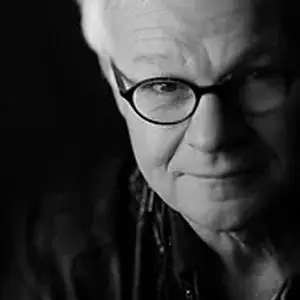 Black and white photo of an older person with short, light hair, wearing glasses. Half of the face is lit, showing a gentle expression, while the other half fades into darkness.