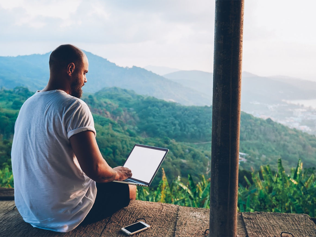 Person working on a laptop in a serene outdoor setting with mountains in the background, representing remote work and inspiration.